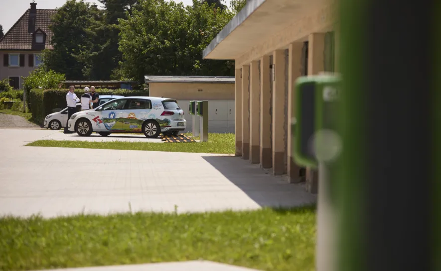 E-Auto an Ladesäule vor einem Wohngebäude mit Personen im Gespräch.